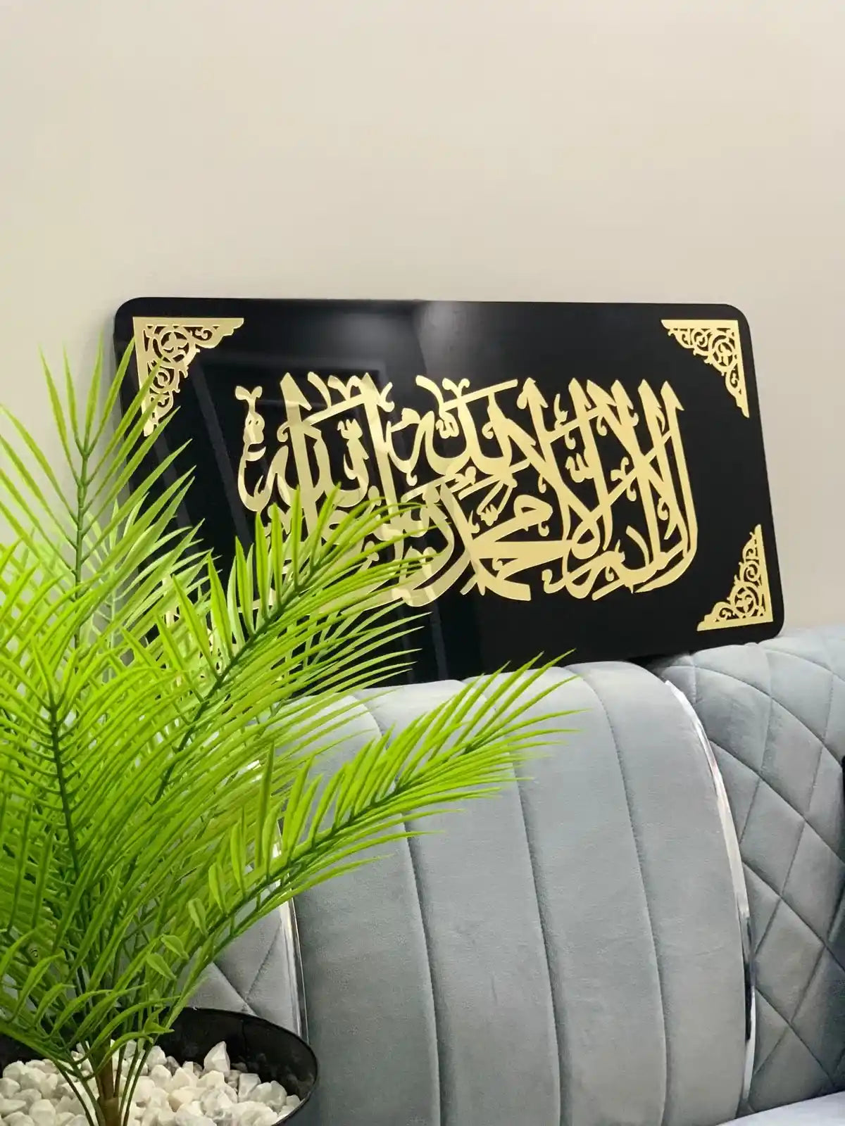 Decorative black plaque with gold Arabic kalma tayyaba calligraphy on a light-colored wall, with a plant in the foreground.