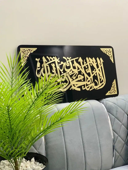 Decorative black plaque with gold Arabic kalma tayyaba calligraphy on a light-colored wall, with a plant in the foreground.